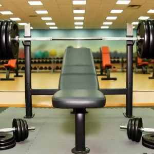 DALL·E 2024-10-19 14.55.38 - A gym setting with a flat bench in the center. On one side of the bench, there is a barbell with plates, and on the other side, there are two heavy du